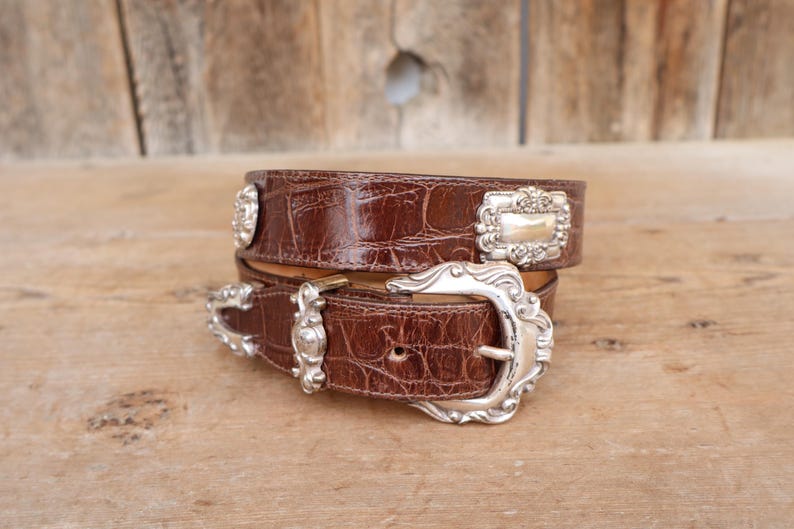 Op de afbeelding: Een bruine leren riem met een krokodillenstructuur en zilverkleurige metalen gesp en accenten. De riem is opgerold en wordt weergegeven op een houten oppervlak. De gesp heeft een sierlijk ontwerp.