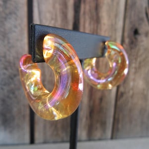 May include: A pair of iridescent gold hoop earrings. The earrings are made of clear plastic with a gold and iridescent finish.