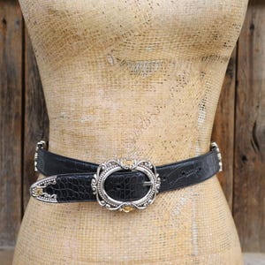 May include: Black leather belt with a silver oval buckle and decorative silver accents. The belt has a crocodile texture and is displayed on a mannequin. The buckle has a silver frame with a gold-colored center.
