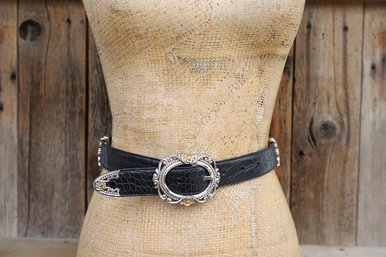 May include: Black leather belt with a silver oval buckle and decorative silver accents. The belt has a crocodile texture and is displayed on a mannequin. The buckle has a silver frame with a gold-colored center.