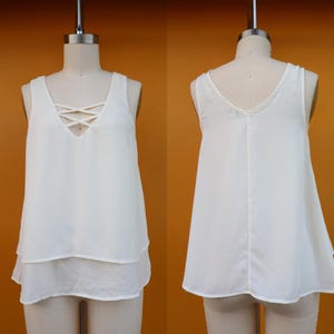 Puede incluir: Top blanco sin mangas con diseño en capas. El top presenta un detalle entrecruzado en el escote y una silueta fluida y en capas. La tela parece ligera. El top se muestra en un maniquí.