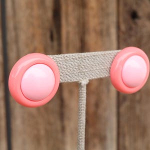 May include: A pair of vintage clip-on earrings. Each earring features a light pink center surrounded by a coral pink border. The earrings are displayed on a neutral-colored earring stand against a wooden background.