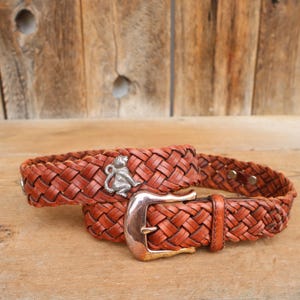 May include: A brown leather belt with a woven design. The belt features a silver buckle and a silver monkey charm. The belt is approximately 3.8 cm wide.