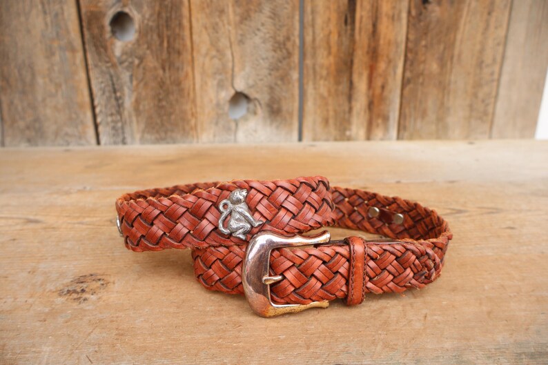 May include: A brown leather belt with a woven design. The belt features a silver buckle and a silver monkey charm. The belt is approximately 3.8 cm wide.