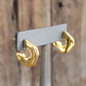 May include: A pair of polished gold-tone hoop earrings featuring a distinct geometric, angular shape. The earrings have a smooth, reflective surface and are designed as open, thick hoops with a post-back closure for secure attachment to the ear.