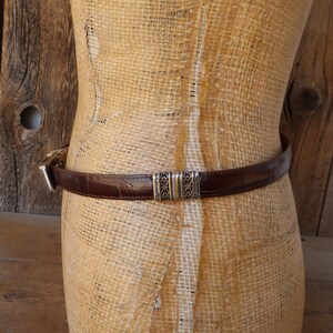 May include: A brown leather belt with a crocodile texture is wrapped around a mannequin. The belt has a silver buckle and a decorative silver center piece. The belt is approximately 1 inch wide.