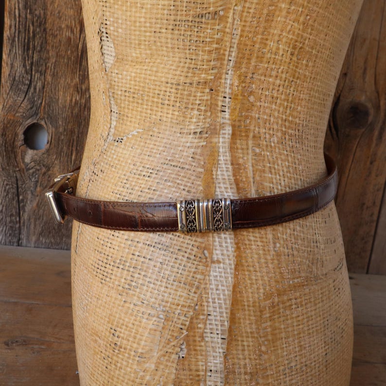 May include: A brown leather belt with a crocodile texture is wrapped around a mannequin. The belt has a silver buckle and a decorative silver center piece. The belt is approximately 1 inch wide.