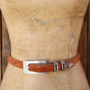May include: A tan leather belt featuring a rectangular silver-tone metal buckle with engraved chevron patterns, two matching silver-tone metal belt loops, and a pointed silver-tone metal tip with a repeating chevron design.