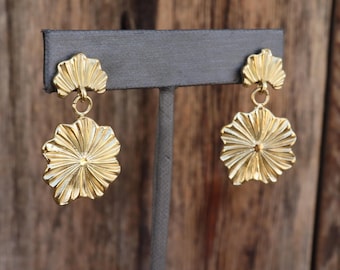 Op de afbeelding: Een paar goudkleurige oorbellen met een bloemmotief. Elke oorbel heeft een kleiner, waaiervormig bovenstuk dat is verbonden met een grotere, bloemvormige hanger. De oorbellen worden getoond op een grijze standaard tegen een houten achtergrond.