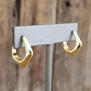 May include: A pair of gold-colored, geometric hoop earrings. The earrings have a squared-off, angular design, with a polished, reflective surface. The earrings are displayed on a gray earring holder against a blurred wooden background.