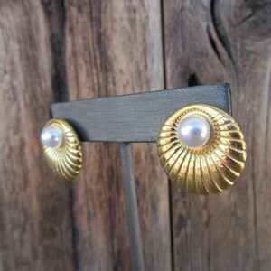 May include: Gold-coloured clip-on earrings featuring a pearl centre and a ridged, spiral design. The earrings are round and have a vintage aesthetic. The earrings are displayed on a grey earring stand against a wooden background.