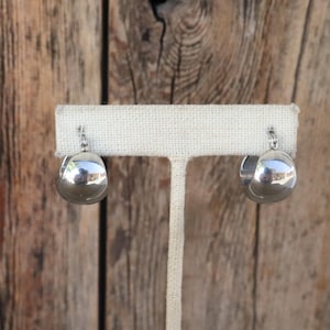 May include: A pair of polished silver hoop earrings. Each earring has a rounded, teardrop shape, reflecting the surrounding environment. The earrings are displayed on a white fabric earring stand against a wooden background.