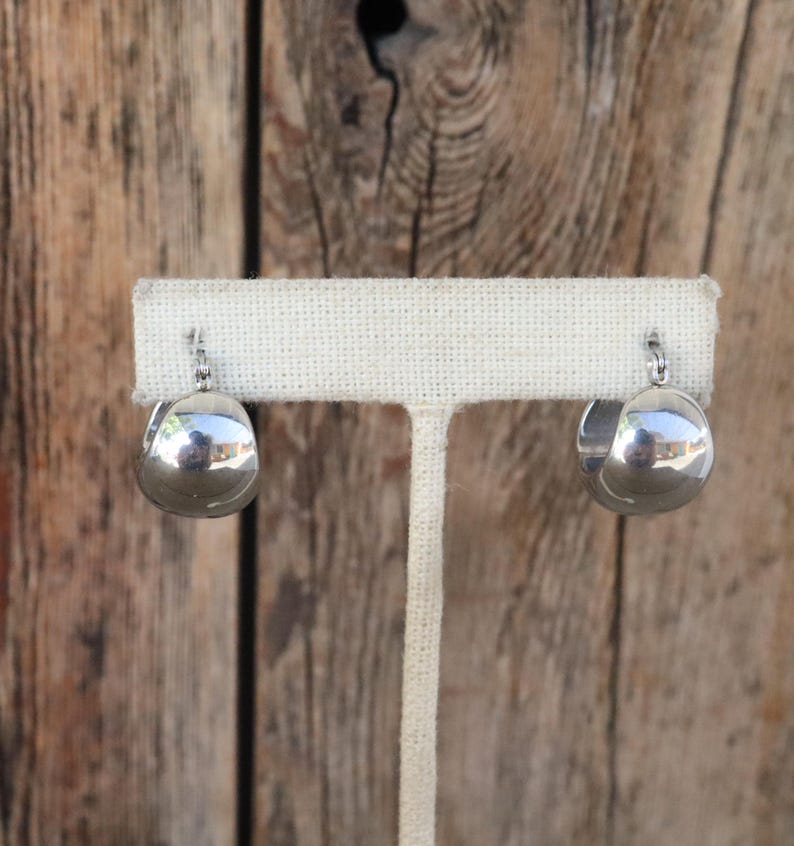 May include: A pair of polished silver hoop earrings. Each earring has a rounded, teardrop shape, reflecting the surrounding environment. The earrings are displayed on a white fabric earring stand against a wooden background.