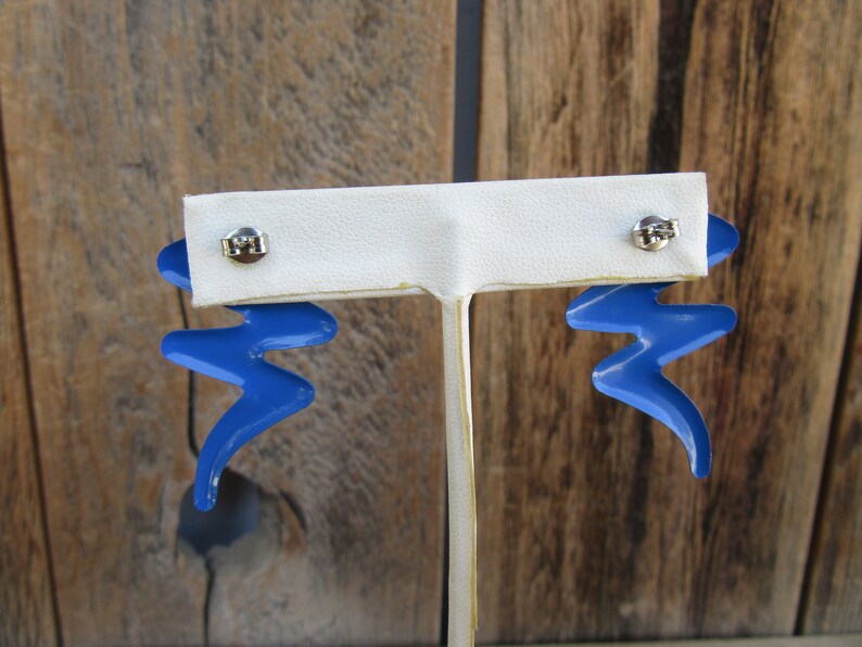May include: A pair of blue lightning bolt shaped earrings with silver posts. The earrings are displayed on a white earring stand.