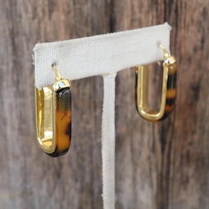 May include: Gold-tone rectangular hoop earrings with a tortoiseshell pattern in brown and black. The earrings are displayed on a white fabric stand. The background is a wooden surface.