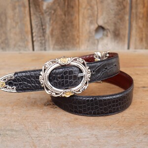 May include: Black leather belt with a silver buckle and gold accents. The belt has a crocodile texture and a brown interior. The buckle is oval-shaped with ornate detailing. The belt is lying on a wooden surface.