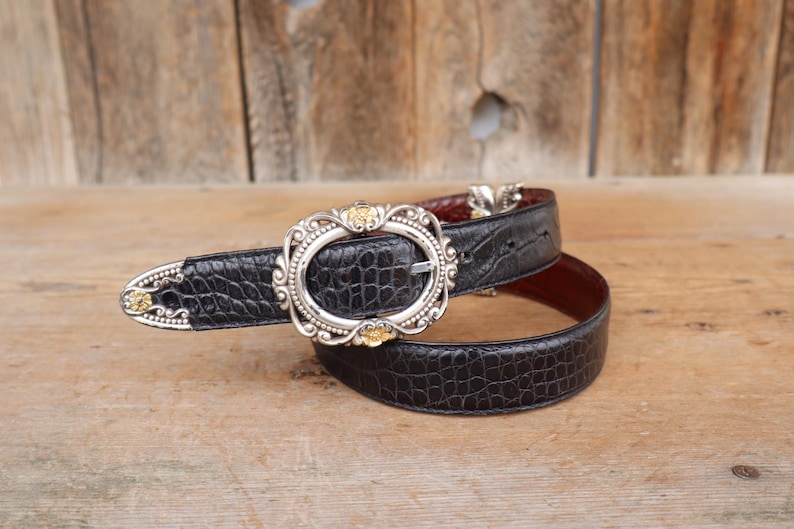 May include: Black leather belt with a silver buckle and gold accents. The belt has a crocodile texture and a brown interior. The buckle is oval-shaped with ornate detailing. The belt is lying on a wooden surface.