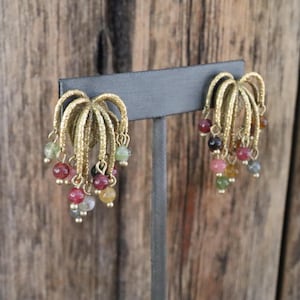 May include: A pair of gold-toned earrings with a textured, fan-like design. Each earring features multiple strands of small, colorful beads in shades of red, green, pink, and yellow, creating a dangling effect. The earrings are displayed against a neutral background.