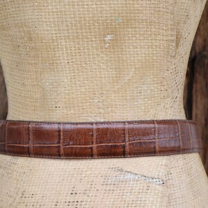 May include: A brown leather belt with a crocodile pattern is displayed on a mannequin. The belt is a medium brown color and has a rectangular pattern. The mannequin is covered in a woven material, and the background is a wooden surface.