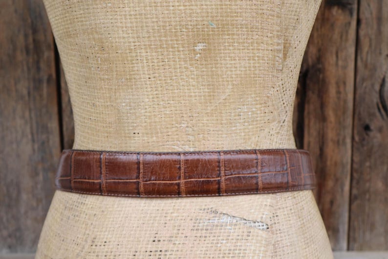 May include: A brown leather belt with a crocodile pattern is displayed on a mannequin. The belt is a medium brown color and has a rectangular pattern. The mannequin is covered in a woven material, and the background is a wooden surface.