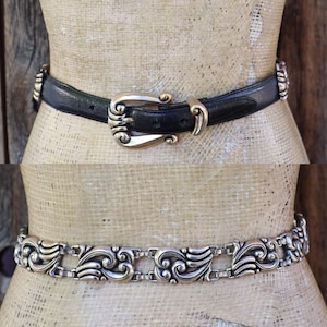 May include: Two belts are displayed on a mannequin. The top belt is black leather with a gold-colored buckle and accents. The bottom belt is silver-toned metal with an ornate, repeating design. Both belts are fashion accessories.