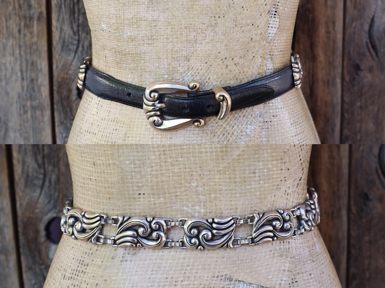 May include: Two belts are displayed on a mannequin. The top belt is black leather with a gold-colored buckle and accents. The bottom belt is silver-toned metal with an ornate, repeating design. Both belts are fashion accessories.