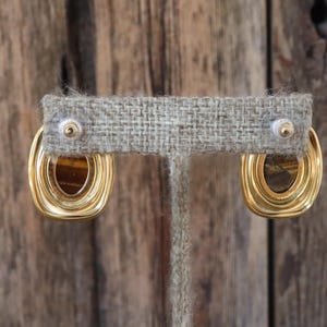 K&ouml;nnte beinhalten: Goldfarbene Ohrringe mit rechteckigem, ovalem Design. Die Ohrringe haben einen geschichteten, strukturierten Goldrahmen, der ein braunes, tigerauge&auml;hnliches Zentrum umgibt. Die Ohrringe werden auf einem neutralfarbenen Ohrringst&auml;nder pr&auml;sentiert.