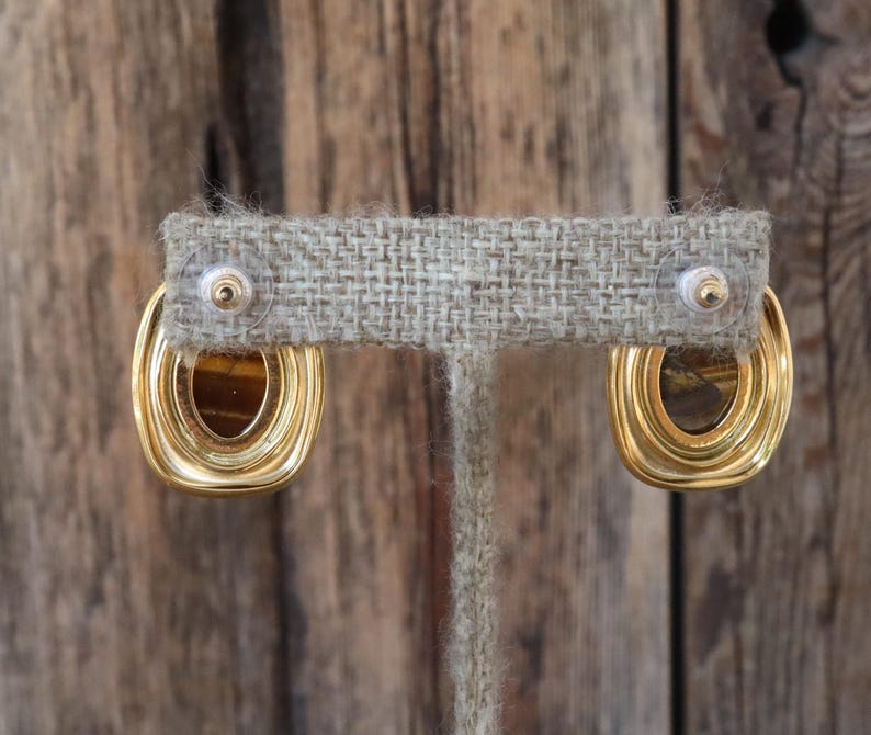 K&ouml;nnte beinhalten: Goldfarbene Ohrringe mit rechteckigem, ovalem Design. Die Ohrringe haben einen geschichteten, strukturierten Goldrahmen, der ein braunes, tigerauge&auml;hnliches Zentrum umgibt. Die Ohrringe werden auf einem neutralfarbenen Ohrringst&auml;nder pr&auml;sentiert.