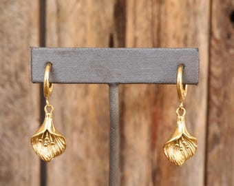Op de afbeelding: Een paar goudkleurige oorringen met een calla lelie ontwerp. De oorbellen hangen aan de ringen en hebben gedetailleerde, getextureerde leliebloesems. De achtergrond is een houten oppervlak.
