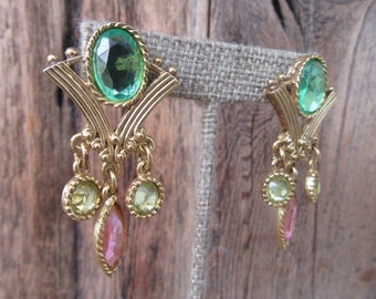 Op de afbeelding: Vintage goudkleurige oorbellen met groene en roze edelstenen. Elke oorbel heeft een grote ovale groene steen aan de bovenkant, met kleinere groene en roze stenen die eronder hangen. De oorbellen hebben een sierlijk, Art Deco-geïnspireerd ontwerp.