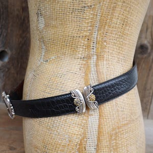 May include: Black leather belt with a textured pattern, featuring ornate silver-tone and gold-tone metal buckle details. The belt is displayed on a mannequin against a rustic wooden background.