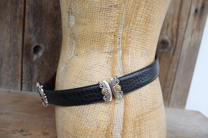 May include: Black leather belt with a textured pattern, featuring ornate silver-tone and gold-tone metal buckle details. The belt is displayed on a mannequin against a rustic wooden background.