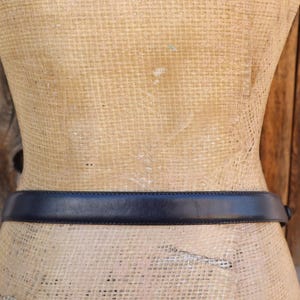 May include: A dark navy blue leather belt is wrapped around a mannequin. The belt is a simple design with a rectangular buckle. The mannequin is tan and the background is a wooden wall.