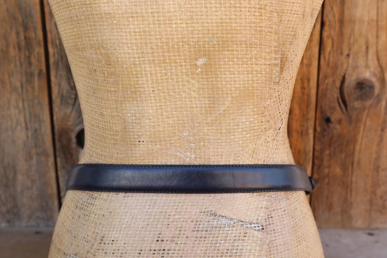 May include: A dark navy blue leather belt is wrapped around a mannequin. The belt is a simple design with a rectangular buckle. The mannequin is tan and the background is a wooden wall.