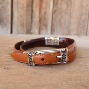 May include: A brown leather belt with a silver buckle and decorative accents. The belt has a light brown exterior and a darker brown interior. The buckle is rectangular with an ornate design. The belt is approximately 1.25 inches wide.