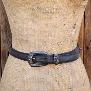 May include: Black leather belt with a silver buckle and decorative accents. The belt is displayed on a mannequin and has a classic western style. The buckle and belt loops have detailed designs.