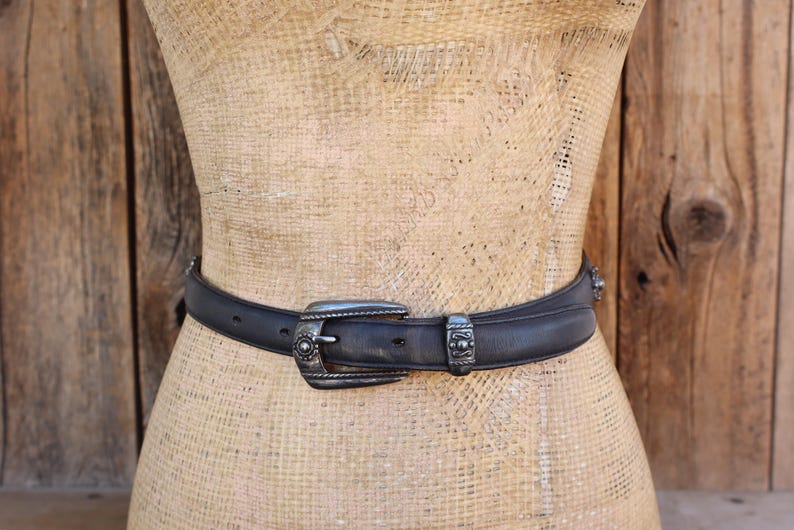 May include: Black leather belt with a silver buckle and decorative accents. The belt is displayed on a mannequin and has a classic western style. The buckle and belt loops have detailed designs.