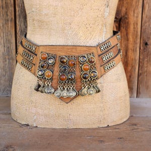 Puede incluir: Un cinturón ancho de cuero marrón con detalles metálicos decorativos y piedras de color ámbar. El cinturón presenta tres secciones con colgantes metálicos y se exhibe en un maniquí.