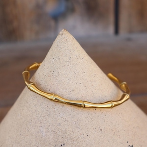 May include: A gold-colored bamboo-style bracelet. The bracelet is made of a shiny, metallic material and is resting on a light-colored, cone-shaped object. The background is a blurred wooden surface.