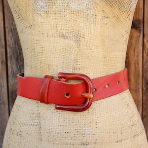 May include: A red leather belt with a dark brown buckle is displayed on a mannequin. The belt has several holes for adjustment and a slightly textured surface. The buckle is a rounded shape.