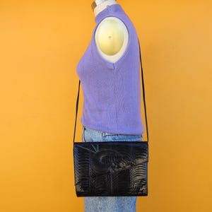 May include: Black rectangular handbag with a textured, embossed pattern. The bag has a long, thin strap and a flap closure. The background is a solid, bright yellow-orange.