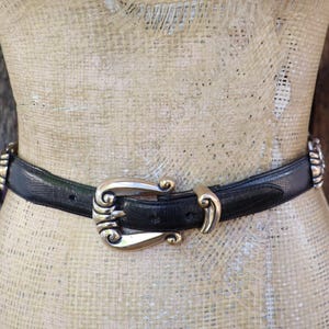 May include: A black belt with a decorative gold-colored buckle and accents. The belt is worn around a mannequin. The buckle has an ornate design. The belt is made of a textured material.