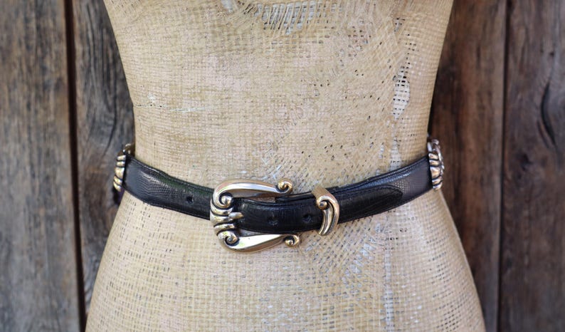 May include: A black belt with a decorative gold-colored buckle and accents. The belt is worn around a mannequin. The buckle has an ornate design. The belt is made of a textured material.