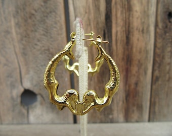 May include: Gold hoop earrings featuring a mermaid design. The mermaids are facing each other and their tails form the hoop.