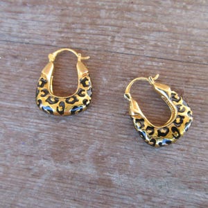 May include: A pair of gold-colored hoop earrings with a leopard print design. The earrings have a rounded, oval shape with a hinged closure. The leopard print features black spots on a tan background.