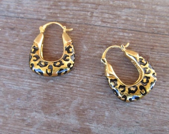 May include: A pair of gold-colored hoop earrings with a leopard print design. The earrings have a rounded, oval shape with a hinged closure. The leopard print features black spots on a tan background.