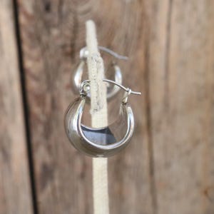 May include: A pair of silver hoop earrings. The earrings are a classic, rounded design with a polished finish. They are displayed on a neutral-colored, textured background.