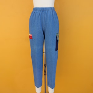 May include: Light blue pants with an elastic waistband and two pockets. The pockets are decorated with colorful fabric patches. The pants are displayed on a mannequin against a yellow background.