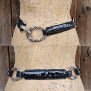 May include: A black belt with a crocodile-textured pattern. The belt features a silver-colored, ornate buckle with a circular design. The belt is displayed on a mannequin against a wooden background.