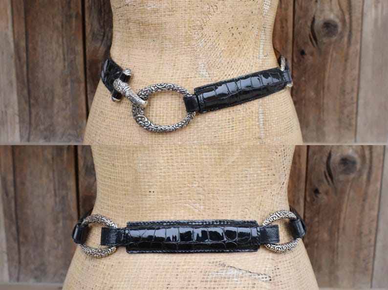 May include: A black belt with a crocodile-textured pattern. The belt features a silver-colored, ornate buckle with a circular design. The belt is displayed on a mannequin against a wooden background.